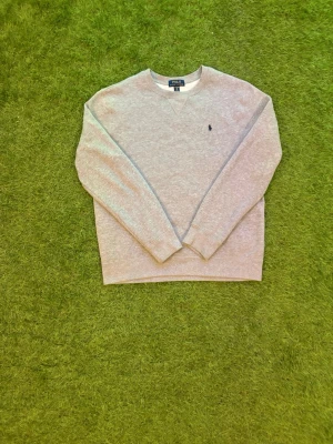 Grå sweatshirt från Polo Ralph Lauren 18/20 år - Klassisk grå sweatshirt från Polo Ralph Lauren med rund halsringning och broderad marinblå logga på bröstet. Tröjan har långa ärmar och ribbade muddar vid ärmslut och nederkant. Perfekt för en avslappnad och stilren look. Passar perfekt om man är 176 typ