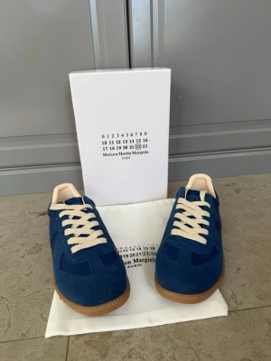 Blå sneakers Maison Margiela - Säljer ett par blå sneakers från Maison Margiela med beige snörning och gummisula. Skorna har en klassisk rund tå och är tillverkade i mocka och skinn för en lyxig känsla. Perfekta för dig som vill ha något stilrent och trendigt.