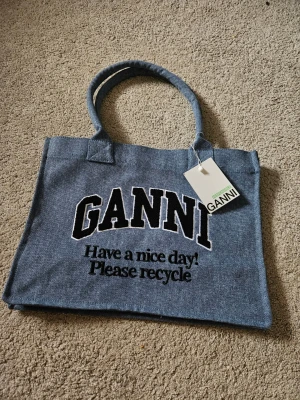 Blå tote bag från Ganni - Trendig blå shoppingväska från Ganni i ett slitstarkt tygmaterial. Väskan har två rejäla handtag och stor broderad logga framtill med texten 'Have a nice day! Please recycle'. Perfekt för att bära dina grejer med stil.