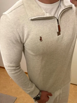 Vit långärmad polotopp med half zip - Snygg vit långärmad polotopp med half zip och ribbad struktur. Toppen har en liten broderad logga på bröstet och en hög krage som kan bäras öppen eller stängd. Perfekt för en clean och stilren look.