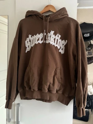 Brun hoodie från Sweet SKTBS - Brun hoodie från Sweet SKTBS med stor vit logotyp på bröstet. Tröjan har en rymlig huva med dragsko, en stor magficka och ribbade muddar vid ärmslut och nederkant. Perfekt för dig som gillar streetwear och avslappnad stil. Den har använts ett fåtal gånger och är i nyskick❤️‍🔥