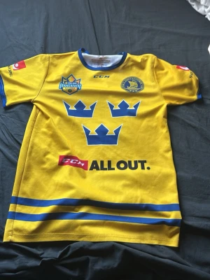 Gul Tre Kronor hockeytröja CCM - Säljer en gul hockeytröja med Tre Kronor-tryck framtill, blå detaljer och loggor på bröstet. Tröjan har korta ärmar, rund halsringning och blå ränder längst ner. Perfekt för dig som gillar hockey och vill stötta Sverige.