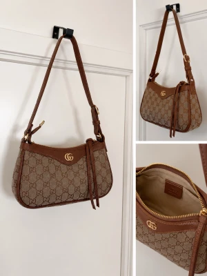 Helt ny axelremsväska - Säljer en snygg axelremsväska  i klassiskt beige och brunt monogramtyg med bruna läderdetaljer. Väskan har guldfärgad logga, justerbar rem och dragkedja upptill. Perfekt för dig som gillar lyxiga accessoarer med ikonisk design.