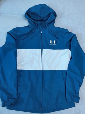 Blå vindjacka från Under Armour - Snygg blå vindjacka från Under Armour med vit bred rand över bröstet och logga på vänster sida. Jackan har huva med dragsko, hel dragkedja och långa ärmar. Perfekt för blåsiga dagar och sportiga outfits.