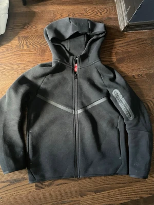 Svart Nike tech hoddie  - Snygg svart Nike tech hoodie med hel dragkedja och huva. Har en diskret grå detalj över bröstet och en praktisk ficka med dragkedja på ena ärmen. Perfekt för en avslappnad och sportig stil.