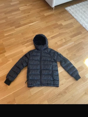 Svart pufferjacka Peak Performance - Snygg svart pufferjacka från Peak Performance med huva och dragkedja framtill. Jackan har quiltad design och långa ärmar med muddar. Perfekt för kalla dagar och enkel att matcha med olika outfits.