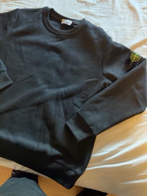 Svart sweatshirt från Stone Island - Säljer en svart sweatshirt från Stone Island med den klassiska loggan på vänster ärm. Tröjan har rund halsringning, ribbade muddar vid ärmslut och nederkant samt en clean och stilren look. Perfekt för dig som gillar streetwear och vill ha något enkelt men ändå ikoniskt.
