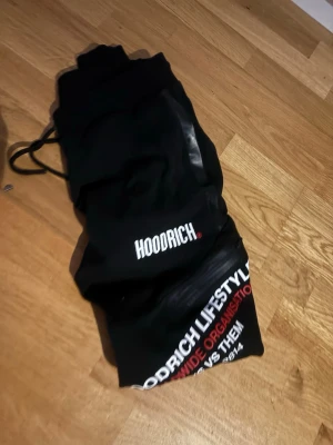 Hoodrich Joggers on Wooden Floor - The images show a pair of black Hoodrich joggers. The first image displays the joggers folded, with the 'HOODRICH' logo visible. The second image shows the joggers laid out flat on a wooden floor, with the text 'HOODRICH LIFESTYLE WORLDWIDE ORGANISATION VS THEM C 2014' printed on the leg in white and red.
