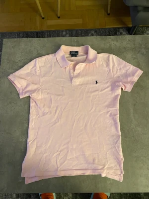 Ljusrosa pikétröja från Ralph Lauren - Klassisk ljusrosa pikétröja från Ralph Lauren med korta ärmar och krage. Tröjan har knappar framtill och den ikoniska broderade loggan på bröstet. Perfekt för en stilren och avslappnad look.