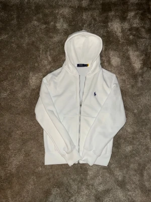 Vit hoodie från Polo Ralph Lauren - Snygg vit hoodie med dragkedja från Polo Ralph Lauren i ny skick. Klassisk design med huva, ribbade muddar och liten broderad logga på bröstet. Perfekt för en clean look nu till hösten. Skickar snabbt📦Kan gå ned i pris vid snabb affär💨