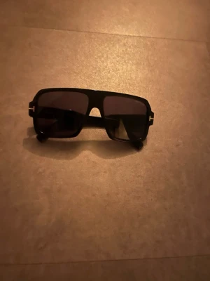 Svarta Tom Ford solglasögon -  Svarta Tom Ford solglasögon med bred rektangulär form och mörka glas. Bågarna är i plast och har en modern, stilren look med diskreta gulddetaljer vid gångjärnen. Perfekta för att höja din outfit under soliga dagar.