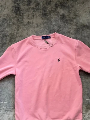 Rosa sweatshirt från Polo Ralph Lauren - Snygg rosa sweatshirt från Polo Ralph Lauren med klassisk rund halsringning och den ikoniska lilla broderade loggan på bröstet. Tröjan har ribbade muddar vid ärmslut och nederkant. Perfekt för en avslappnad och trendig look.