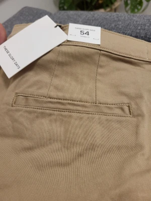 Beige chinos från These Glory Days - Snygga beige chinos från These Glory Days i storlek 54. Klassisk rak passform med fickor fram och bak, bälteshällor och knappgylf. Tillverkade i mjuk bomull för en bekväm känsla och stilren look.