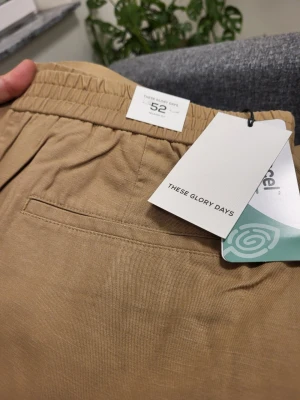Beige shorts från These Glory Days - Snygga beige shorts från These Glory Days med elastisk midja och en bakficka. Tillverkade i ett mjukt bomullsmaterial som passar perfekt till varmare dagar. Enkel och stilren design som är lätt att matcha.