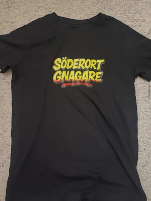 Svart t-shirt med Söderort Gnagare-tryck - Cool svart t-shirt med färgglatt tryck där det står 'Söderort Gnagare' i gult och rött på bröstet. Klassisk passform och rund halsringning, perfekt för dig som vill visa ditt stöd för Söderort.