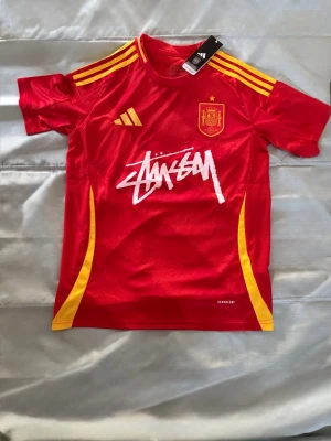 Röd Spanien fotbollströja Adidas - Röd fotbollströja från Adidas med Spaniens landslagsmärke på bröstet, gula detaljer på axlar och sidor samt vit Stüssy-logga framtill. Tillverkad i lätt och ventilerande material, perfekt för träning eller match.