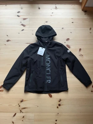 Svart moncler windbreaker  - Snygg svart vindjacka från Moncler med huva och dragkedja framtill. Jackan har stora Moncler-trycket på framsidan, två dragkedjefickor och justerbara muddar. Materialet är lätt och vattenavvisande, perfekt för blåsiga dagar. Klassisk Moncler-logga på ärmen. Den är helt ny och aldrig använd. Riktigt fett jacka nu till hösten! 