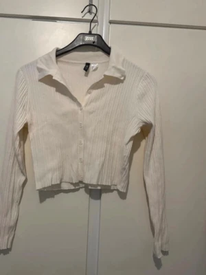 Vit ribbad cropped cardigan - Säljer en vit ribbad cardigan med knappar framtill och krage. Tröjan är cropped och har långa ärmar. Perfekt att styla med höga jeans eller kjol för en clean look. Använd 1 gång 