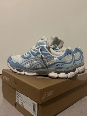  Nya Asics blå och vita sneakers - Snygga sneakers från Asics i vitt och ljusblått med mesh- och syntetmaterial. Skorna har en sportig design med vita snören och en robust, vit sula. Perfekta för dig som gillar en fräsch och trendig look.