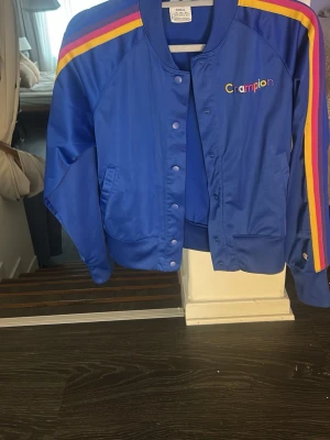 Blå bomberjacka från Champion - Blå bomberjacka från Champion med färgglada ränder i gult, rosa och rött längs axlarna. Jackan har tryckknappar, ribbade muddar och två fickor framtill. Champion-loggan är broderad på bröstet i flera färger. Perfekt för en sportig och trendig look.
