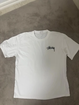 Vit Stüssy t-shirt med tryck - Vit t-shirt från Stüssy med svart logga på bröstet och stort tryck på ryggen med två rosa tärningar och Stüssy-loggan ovanför. Klassisk passform och rund halsringning. Perfekt för dig som gillar streetwear och vill sticka ut.