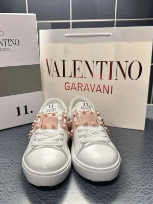 Valentino vita sneakers med nitar - Säljer ett par vita sneakers från Valentino Garavani med ljusrosa band och coola nitar på sidorna. Skorna har en chunky sula och är tillverkade i slätt läder. Perfekta för dig som vill sticka ut med en lyxig och edgy stil.