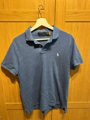 Blå pikétröja från Polo Ralph Lauren - Säljer en blå pikétröja från Polo Ralph Lauren i custom slim fit. Tröjan har korta ärmar, klassisk krage och knappar vid halsen. Extremt sparsamt använd, bara att skriva vid frågor