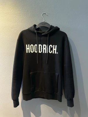 Svart hoodie från Hoodrich - Svart hoodie från Hoodrich med stor vit logga på bröstet. Klassisk huva med dragsnören och en stor magficka framtill. Tillverkad i mjukt material som känns skönt mot huden. Perfekt för en avslappnad och streetinspirerad stil.