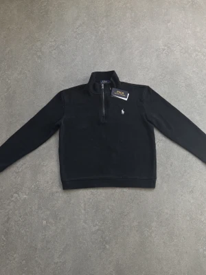 Svart half-zip sweatshirt Polo Ralph Lauren - Klassisk svart sweatshirt från Polo Ralph Lauren med half-zip dragkedja och ståkrage. Tröjan har lång ärm och den ikoniska broderade loggan på bröstet. Tillverkad i mjukt material som passar perfekt till en avslappnad stil. Storlek: Barn L (motsvarar ungefär S/ i vuxenstorlek)