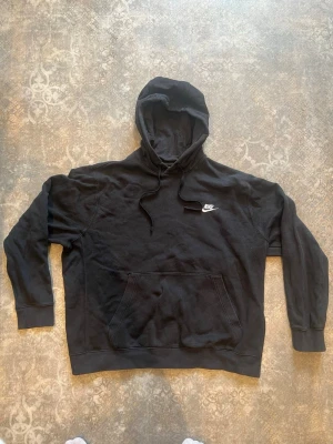 Svart hoodie från Nike  - Säljer en svart hoodie från Nike med klassisk vit logga på bröstet. Hoodien har huva med dragsko och en stor magficka framtill. Tillverkad i mjukt bomullsmaterial och har ribbade muddar vid ärmar och nederkant. Perfekt för en avslappnad och sportig stil.