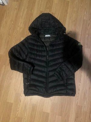 Svart dunjacka med huva - Snygg svart dunjacka med huva och dragkedja framtill. Jackan har två sidofickor med dragkedja och quiltad design. Perfekt för kalla vinterdagar och har en patch på ärmen för extra stil. Obs vänstra fickans dragkedja är sönder