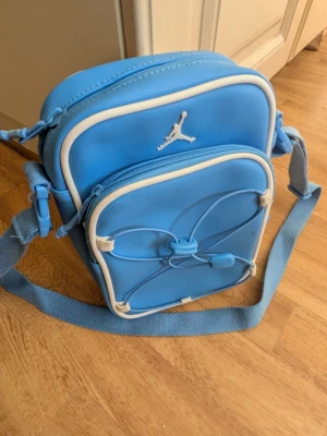 Blå Jordan axelväska med vita detaljer - Snygg blå axelväska från Jordan med vita detaljer och justerbar axelrem. Väskan har två fack med dragkedja, en dekorativ snörning på framsidan och det klassiska Jumpman-loggan. Perfekt för att bära det viktigaste och ger en sportig vibe till din outfit.