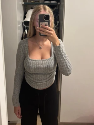 Grå ribbstickad topp med fyrkantig ringning - Trendig grå ribbstickad topp med lång ärm och snygg fyrkantig ringning. Figurnära passform och mjukt material som ger en bekväm känsla. Perfekt att matcha med både jeans och kjol för en stilren look.