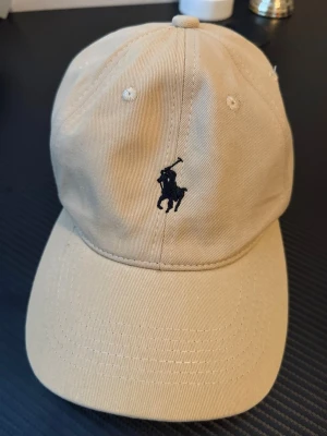 Beige keps från Polo Ralph Lauren - Snygg beige keps från Polo Ralph Lauren med klassisk mörkblå broderad logga framtill. Justerbar rem baktill i metall och bomullsmaterial som ger en skön och avslappnad look. Perfekt accessoar för en stilren och sportig outfit.