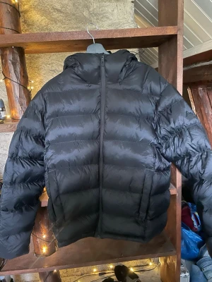 Svart pufferjacka med huva M - Säljer en svart pufferjacka i storlek medium med huva och dragkedja framtill. Jackan har en klassisk quiltad design och är perfekt för kalla vinterdagar. Materialet är glansigt och ger en modern look. Passar dig som vill ha en varm och stilren jacka.