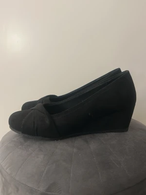 Svarta kilklackade pumps från Graceland - Snygga svarta pumps från Graceland med kilklack och rundad tå. Skorna har en elegant omlott-detalj framtill och är tillverkade i ett mockaliknande material. 