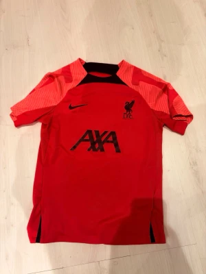 Liverpool tröja  - Säljer en röd Liverpool FC tränings t-shirt från Nike med svarta detaljer och AXA-logga på bröstet. Tröjan har korta ärmar och klubbmärke på vänster sida. Perfekt för fotbollsträning eller sportiga aktiviteter. Mellan storlek XXS-XS
