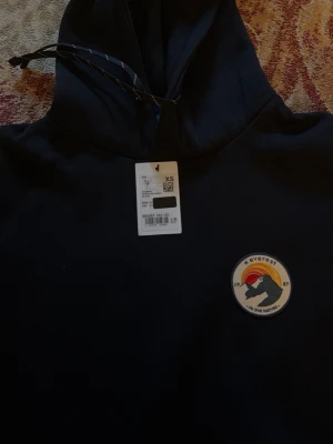 Mörkblå hoodie från Everest XS - Svart hoodie från Everest med broderad patch på bröstet och klassisk känguruficka. Tröjan har huva med dragsko och är tillverkad i mjukt material som passar perfekt för en avslappnad stil.