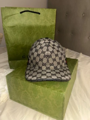 Gucci keps - Snygg keps från Gucci i beige och brunt med det klassiska GG-monogrammönstret över hela kepsen. På sidan finns en röd och marinblå rand. Kepsen har böjd skärm och justerbar rem bak. Materialet är canvas med detaljer i läder.