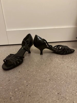 Svarta pumps med remmar från Tesori - Snygga svarta pumps från Tesori med låg klack och öppna tår. Skorna har flera smala remmar över foten och en elegant design som ger en stilren look. Perfekta för dig som gillar klassiska och trendiga skor med en twist.passar perfekt till fest, bröllop, student, midsommar eller en fin middag. Skorna är använda men i fint begagnat skick med normalt slitage på sulorna (se bilder). Bekväma att gå i tack vare den lägre klacken och enkla att matcha med både klänning, kjol och kostymbyxor. 