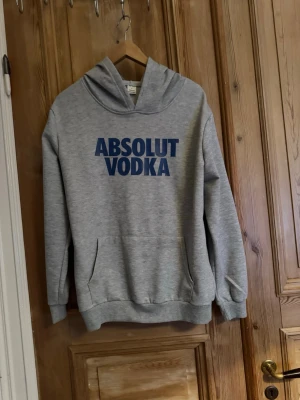 Grå hoodie med ABSOLUT VODKA-tryck - Snygg grå hoodie med blått ABSOLUT VODKA-tryck på bröstet. Klassisk modell med huva och känguruficka framtill. Perfekt för en avslappnad streetstyle-look. Mjuk och bekväm bomullsblandning.