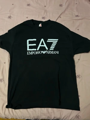Svart EA7 Emporio Armani t-shirt - Storlek L. Snygg svart t-shirt från EA7 Emporio Armani med stor vit logga på bröstet. Klassisk passform och rund halsringning, perfekt till jeans eller shorts. Tillverkad i mjuk bomull för skön känsla hela dagen.