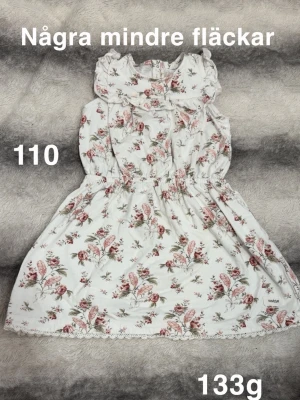 Vit blommig klänning med volang - Söt vit klänning med rosa och grönt blommönster, volang vid halsen och ärmlös design. Resår i midjan för bekväm passform och dekorativ spetskant längst ner. Perfekt för varma dagar och somriga tillfällen.