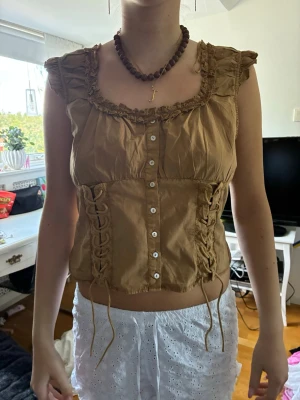 Brun linnetopp med snörning - Snygg brun linnetopp med volangkant vid halsringningen och knappar framtill. Toppen har snörningsdetaljer på sidorna och är ärmlös, vilket ger en trendig och avslappnad look. Perfekt för dig som gillar unika detaljer och en bohemisk stil.