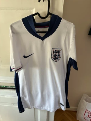 England Nike fotbollströja vit/blå - Snygg England fotbollströja från Nike i vitt med mörkblå detaljer och krage. Tröjan har broderat landslagsemblem på bröstet och Nike-logga. Tillverkad i lätt och ventilerande funktionsmaterial, perfekt för träning eller match.
