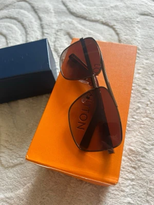 Bruna aviator solglasögon Louis Vuitton - Snygga aviator solglasögon från Louis Vuitton med bruna glas och tunna svarta bågar. Glasen har en lätt toning och loggan syns diskret på sidan. Perfekta för dig som vill ha en klassisk men trendig look. Helt nya sällsynta och oanvända.