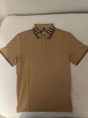 burberry pike - Stilren beige pikétröja med klassisk rutig krage och knappslå framtill. Kragen har svart, vit och röd rutdetalj som ger en snygg kontrast. Kortärmad modell i mjukt material, perfekt för en avslappnad men ändå uppklädd look.