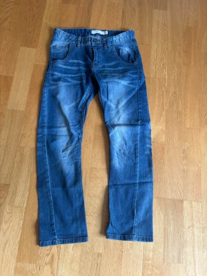Blå  jeans från Lindex - Säljer ett par blå jeans med raka ben och coola slitningar på framsidan och baksidan. Jeansen har klassiska fem fickor och knappgylf. Perfekta för en avslappnad och trendig look.