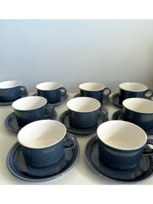 Bo Fajans - 18 delar set med blå koppar och fat - Ikoniska och bejublade servisen med blå / mörkblå kaffekoppar och  fat från anrika svenska keramiktillverkaren Bo Fajans. Tidlösa populära modellen 823 i mörkblå, rustik glasyr med vit insida och en dekorativ flätad kant på faten. Kopparna är cirka 6 cm höga med en diameter på 10 cm, och faten har en diameter på 15 cm. 18-delars set med 9 koppar och 9 fat. Ett stycke svensk designhistoria i keramikform. Mycket gott skick!