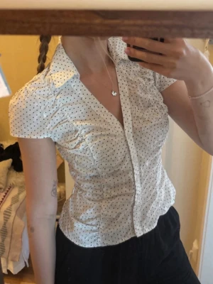 Vit prickig blus  - Säljer en vit blus med svarta små prickar och korta puffärmar. Blusen har en figurnära passform, klassisk krage och knäppning framtill. Materialet känns lätt och luftigt, perfekt för en trendig och stilren look.